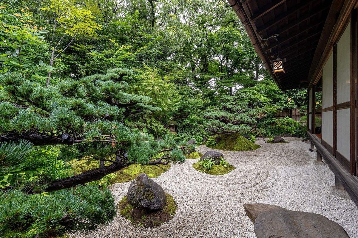 Um jardim japonês tradicional com caminho de cascalho, pinheiros, pedras e um edifício de madeira.