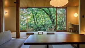 A room with tatami flooring, a low table, a sofa, and a view of a lush green forest through large windows. - Jinya Ryokan (Hadano)