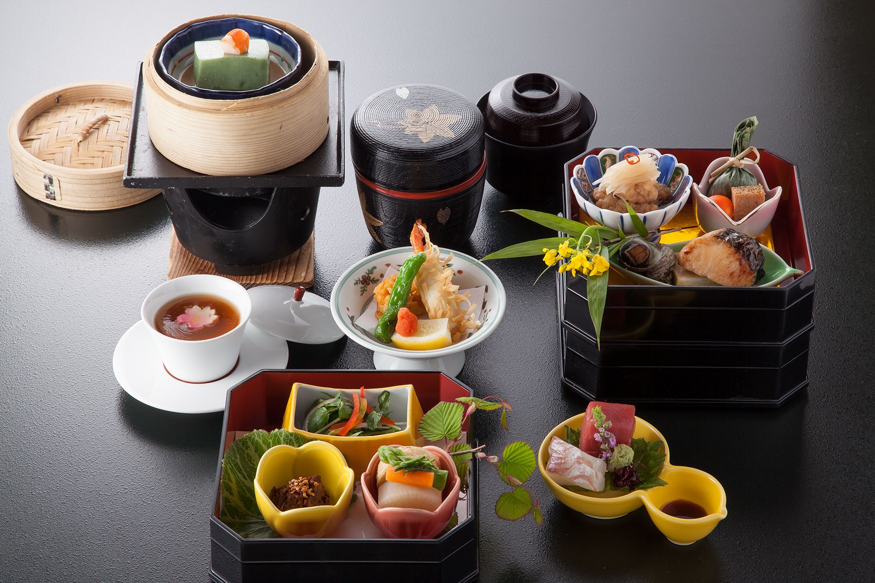 A variety of traditional Japanese dishes and utensils, including a bamboo steamer, a black lacquered container, and several small bowls with different foods.