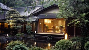 A traditional Japanese-style building with a koi pond in front, wooden deck, and a small outdoor seating area with tables and chairs. - Jinya Ryokan (Hadano)