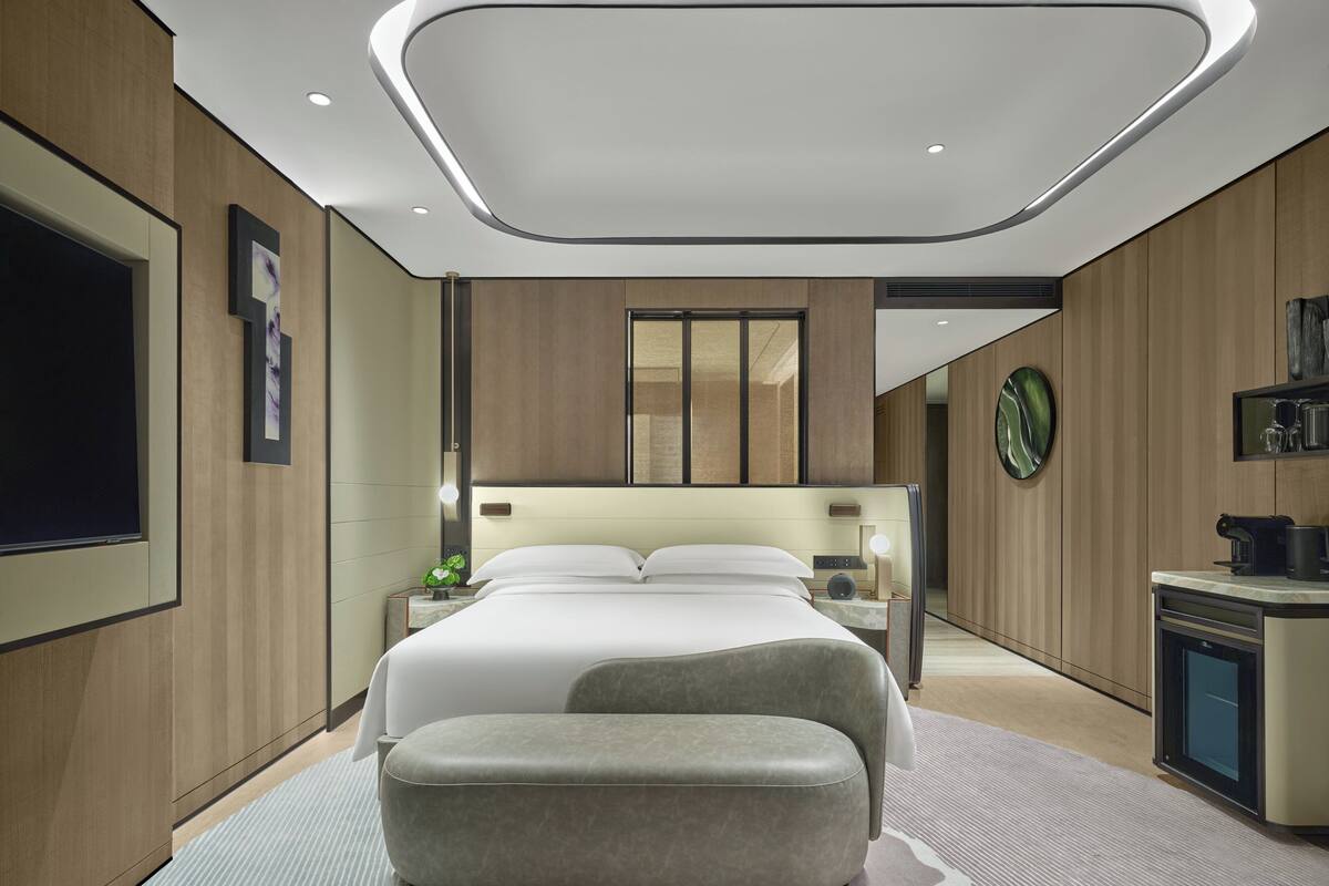 A modern hotel room with a large bed, a seating area, a desk with a coffee machine, and a TV.