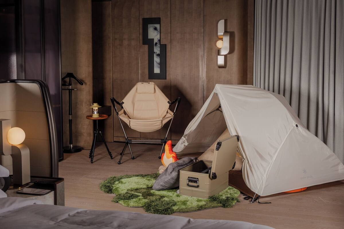 A modern hotel room with a bed, a hanging chair, a tent, and a small table with a lamp.