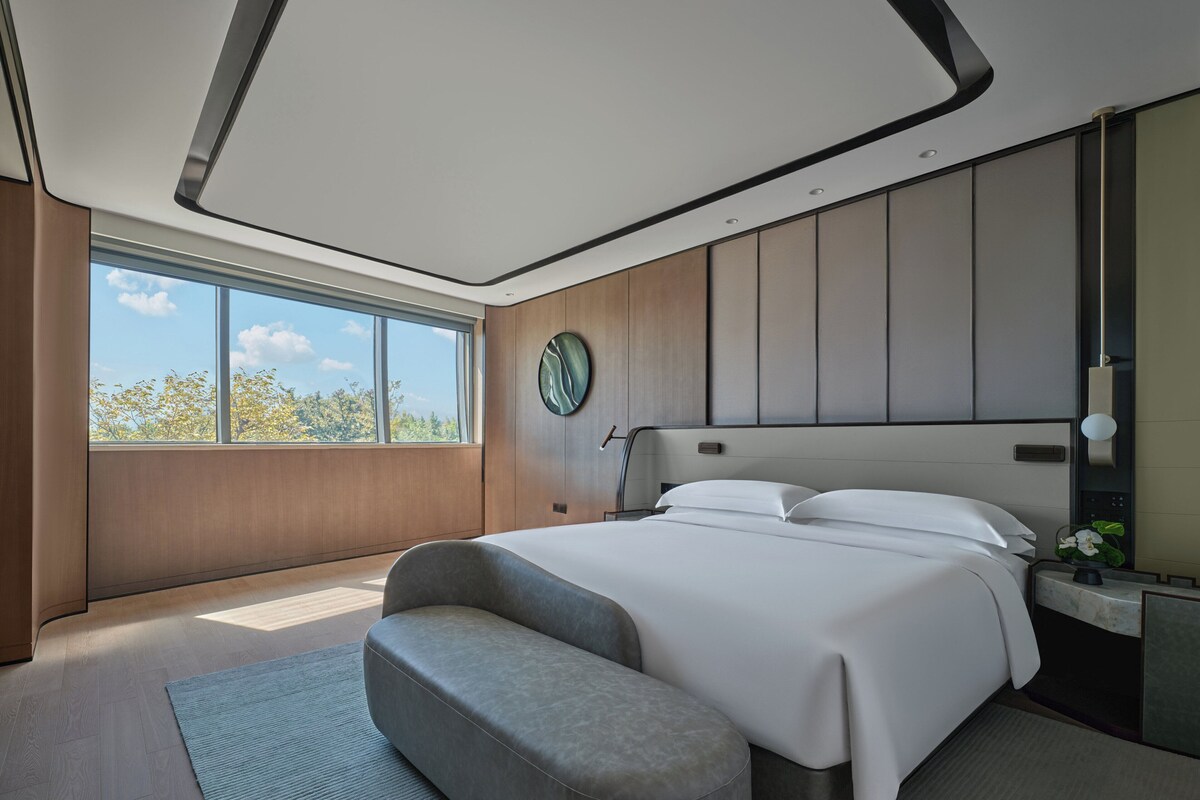 Una habitación de hotel moderna con una cama grande, un área de descanso, un ventanal amplio y vista a árboles.