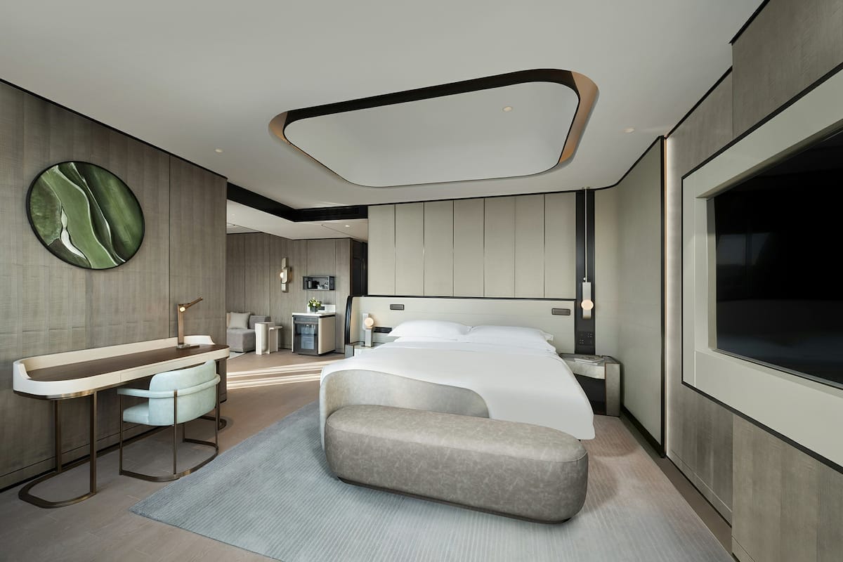 A modern hotel room with a large bed, a sitting area, a vanity, and a large window.
