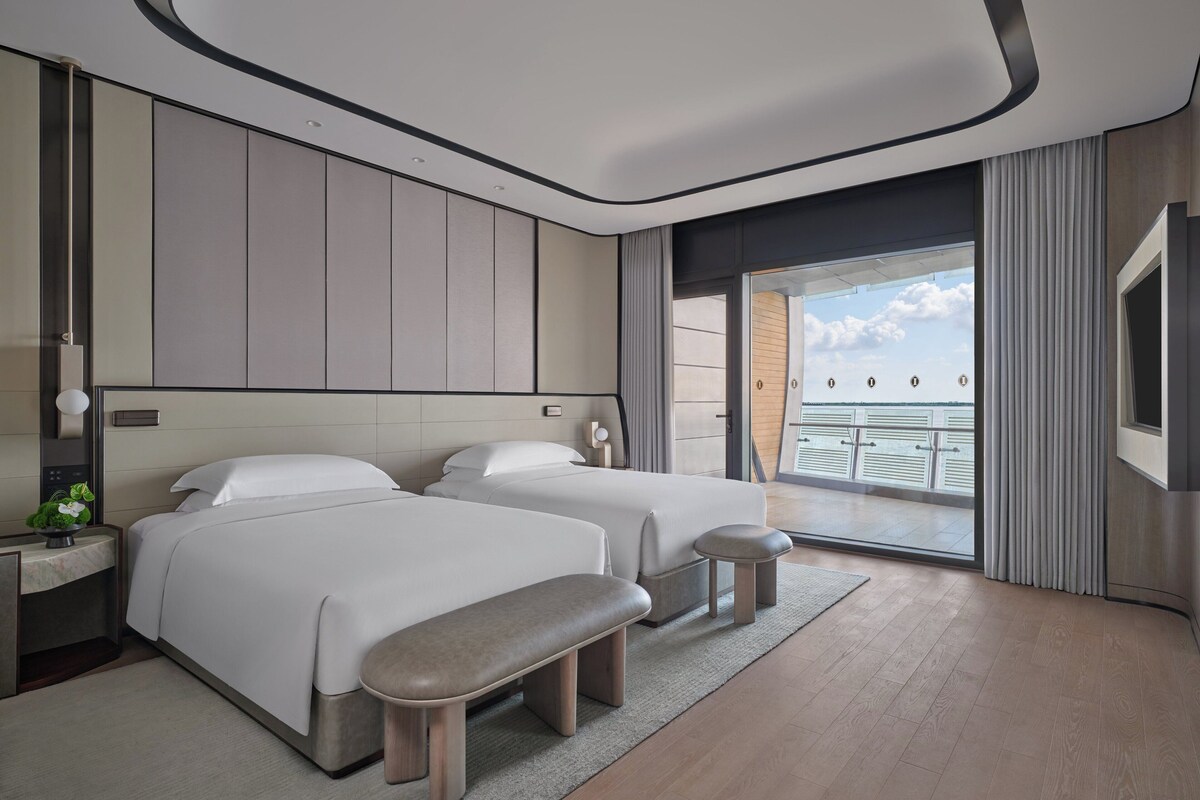 Una habitación de hotel moderna con dos camas, un banco, un balcón con vistas y un ventanal grande.