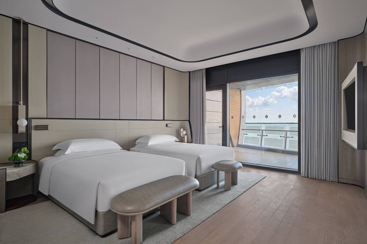 A modern hotel room with two beds, a bench, a balcony with a view, and a large window.