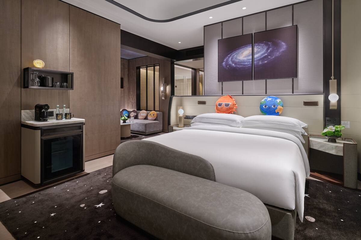 A modern hotel room with a large bed, a sofa, a coffee station, and a wall-mounted TV displaying a galaxy image.