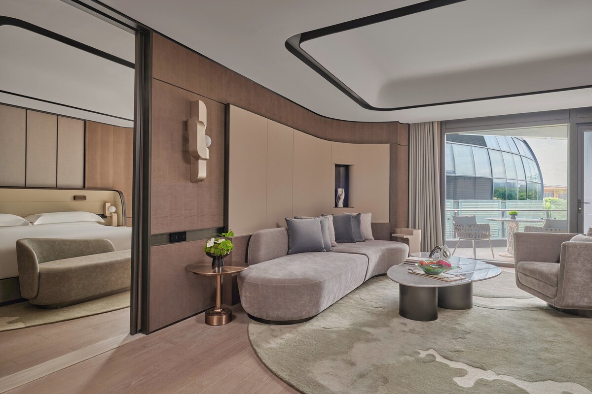 Habitación de hotel moderna con un sofá curvo, una mesa de centro redonda y un amplio ventanal con vistas al exterior.