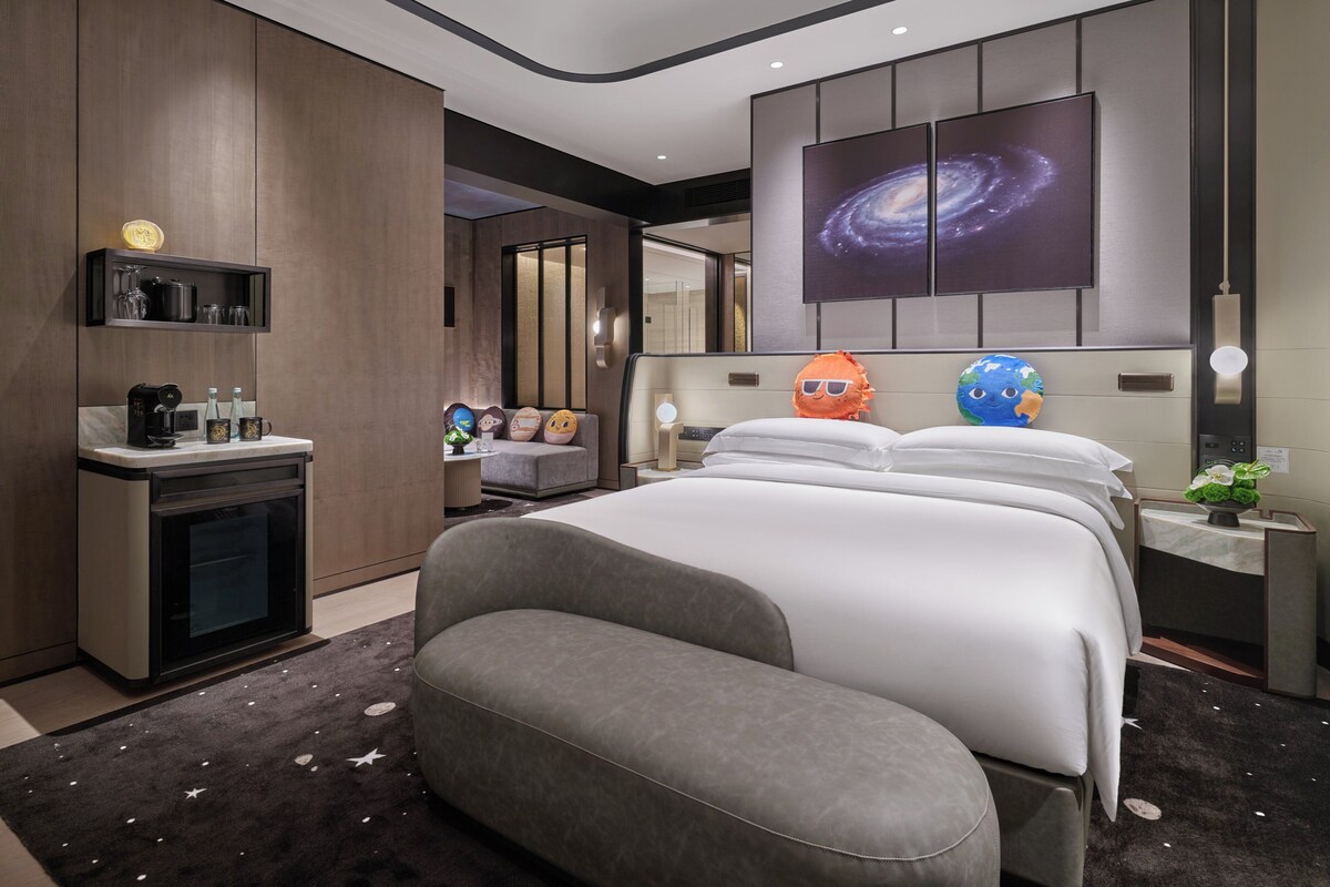 Una habitación de hotel moderna con una cama grande, un sofá, una estación de café y un televisor empotrado mostrando una imagen de una galaxia.