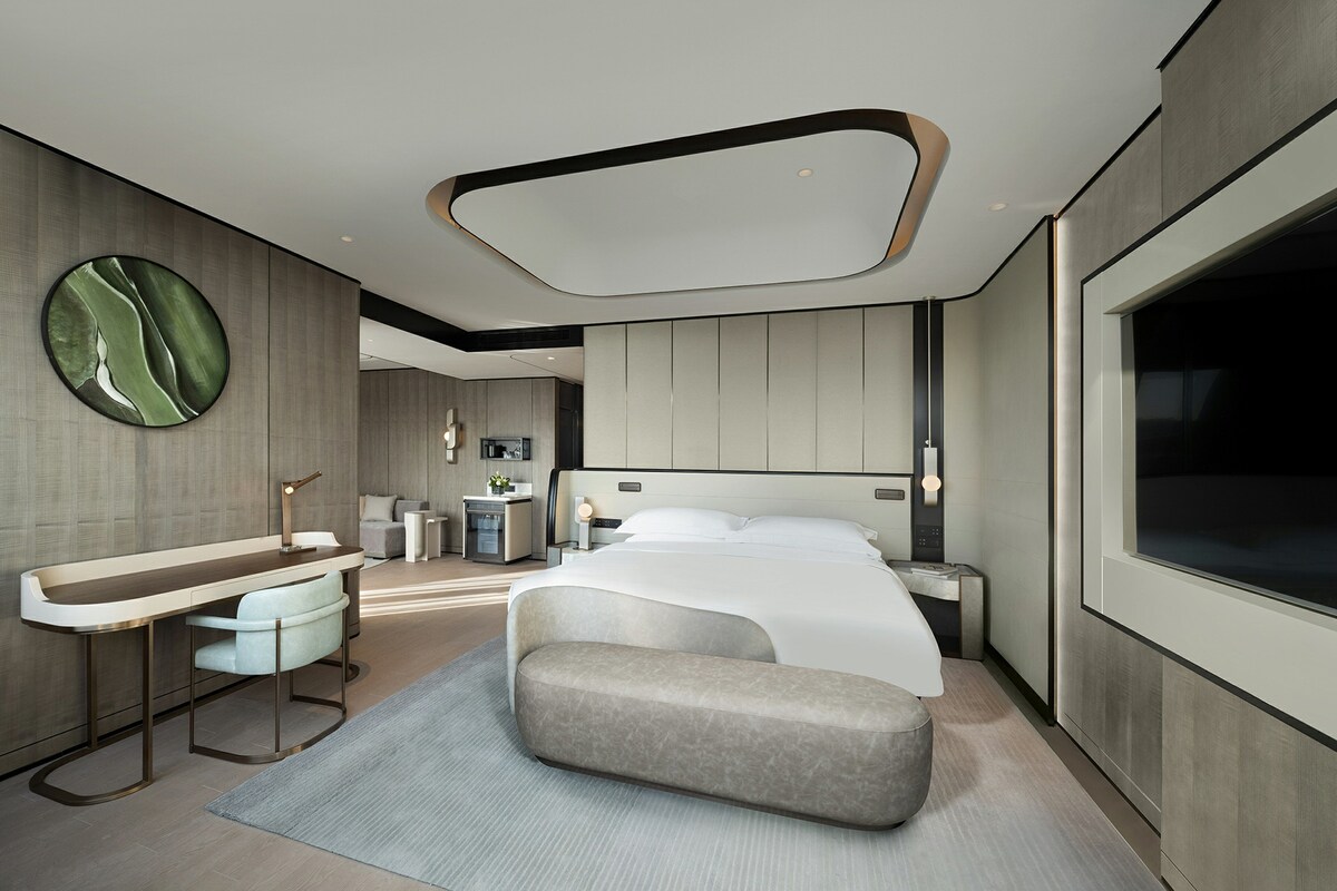 Una habitación de hotel moderna con una cama grande, una zona de estar, un tocador y un ventanal amplio.