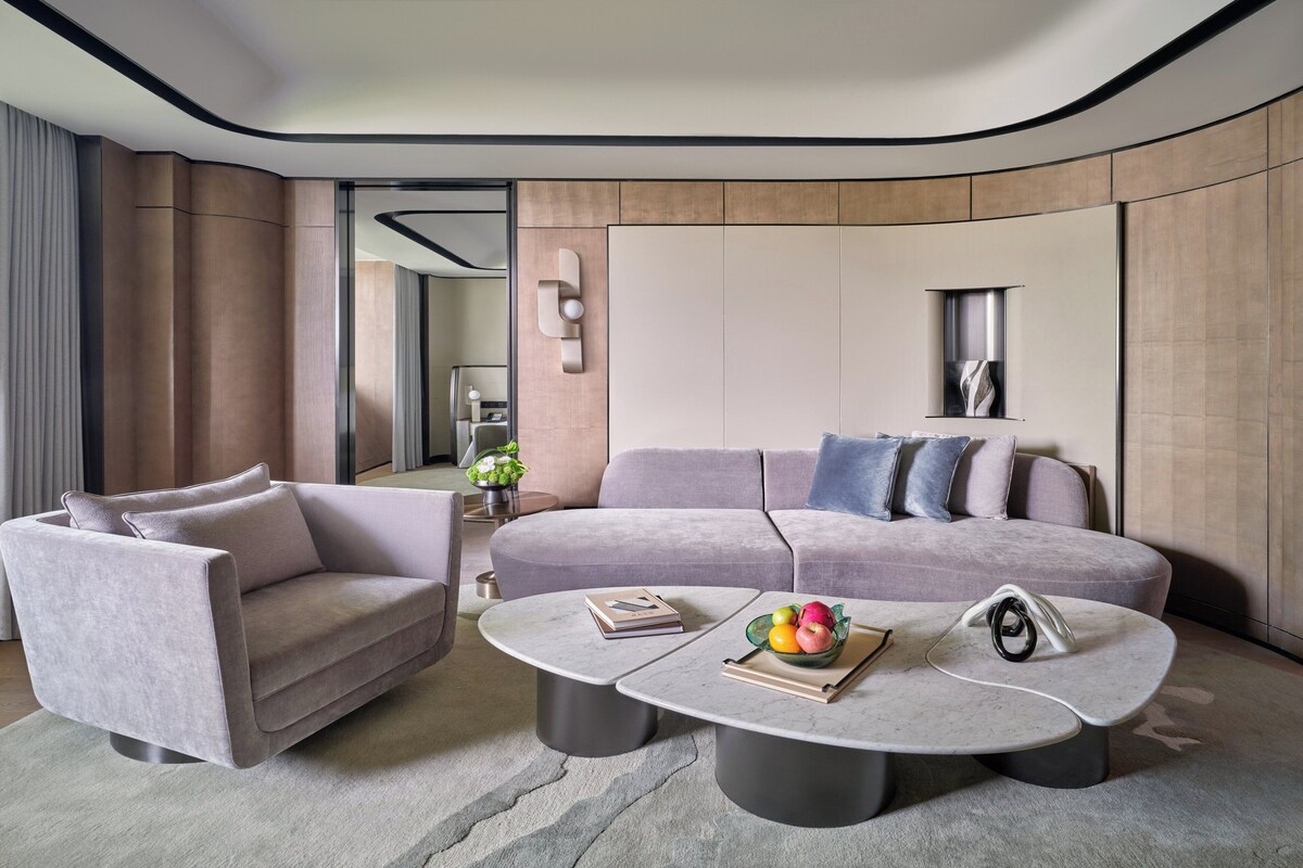 Una sala de estar moderna con un sofá gris, un sillón más pequeño y una mesita de centro con libros y un frutero.
