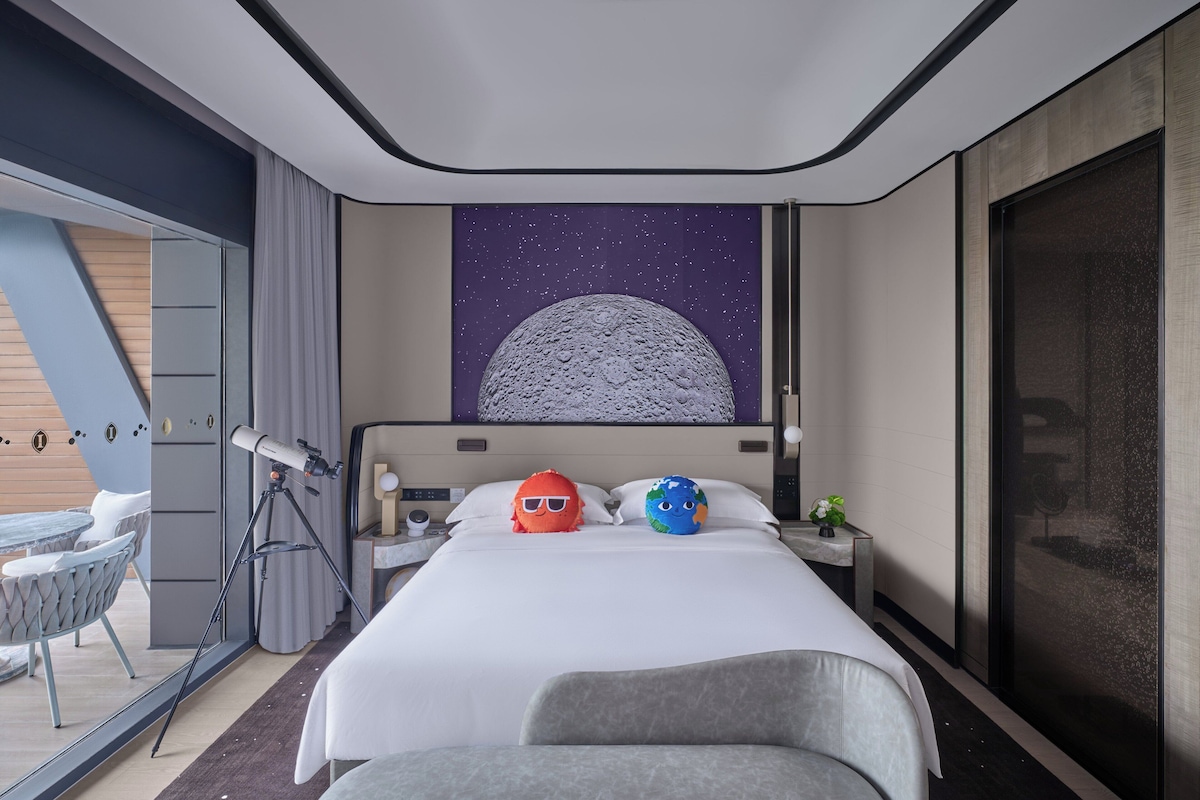 Une chambre d’hôtel moderne avec un grand lit, un télescope et une fresque représentant la lune.