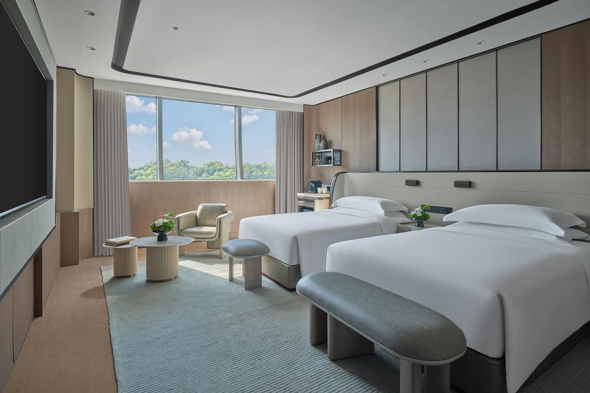 A modern hotel room with two beds, a seating area, a large window, and a view of the outdoors.