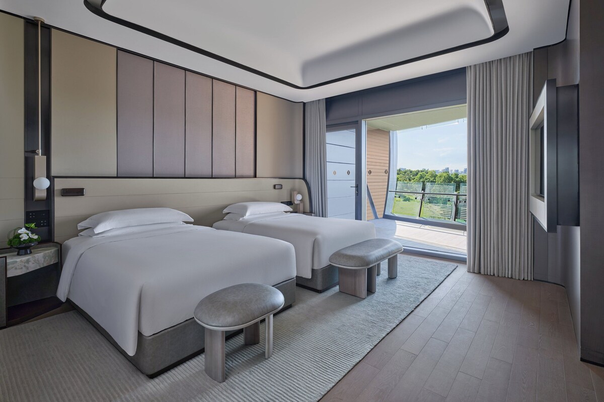 Una habitación de hotel moderna con dos camas, un taburete pequeño, una mesita de noche y un ventanal grande con vistas al exterior.