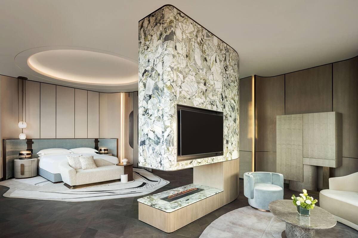 A modern hotel room with a large TV mounted on a marble wall, a sofa, a coffee table, and a round rug.