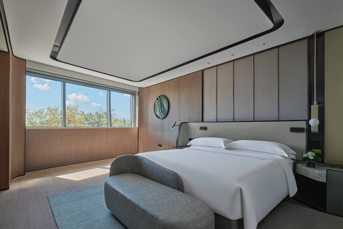 A modern hotel room with a large bed, a seating area, a large window, and a view of trees.