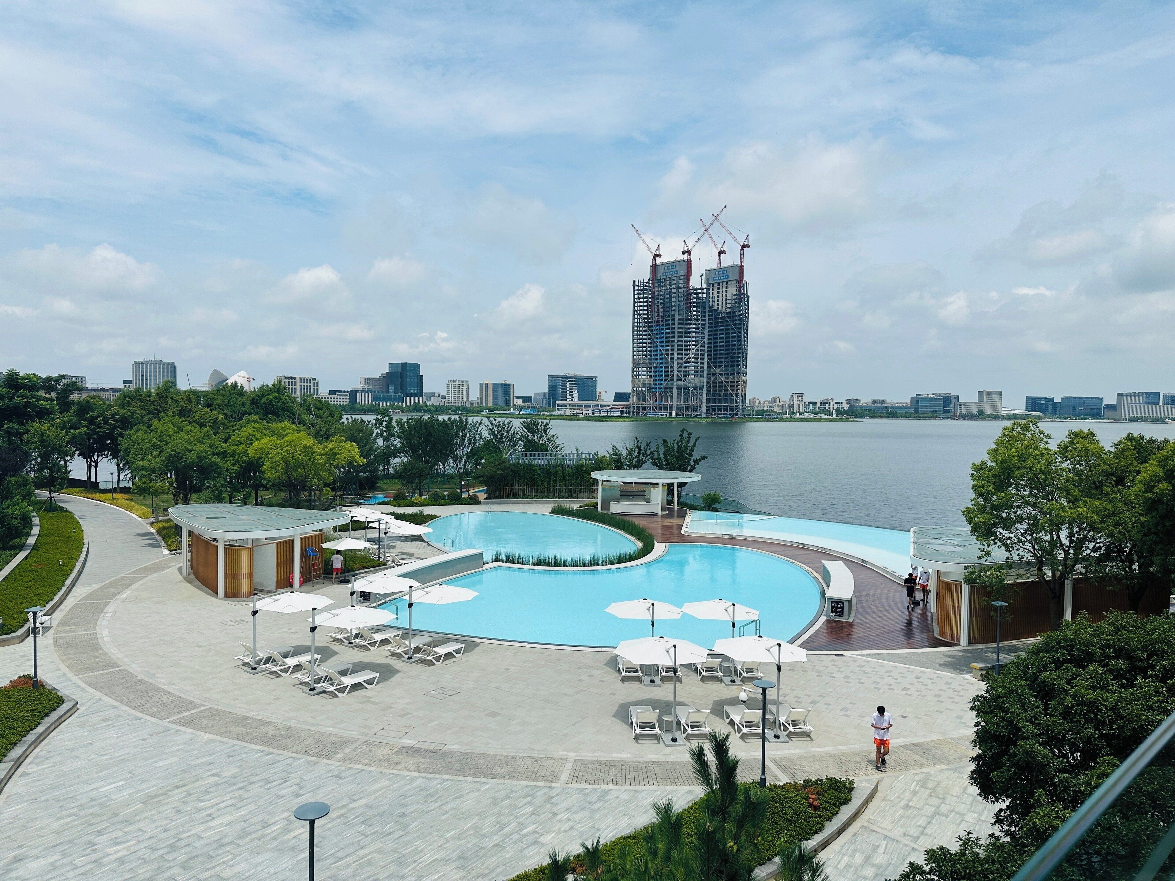 A swimming pool area with lounge chairs and umbrellas, surrounded by greenery and a view of a city skyline with a building under construction.