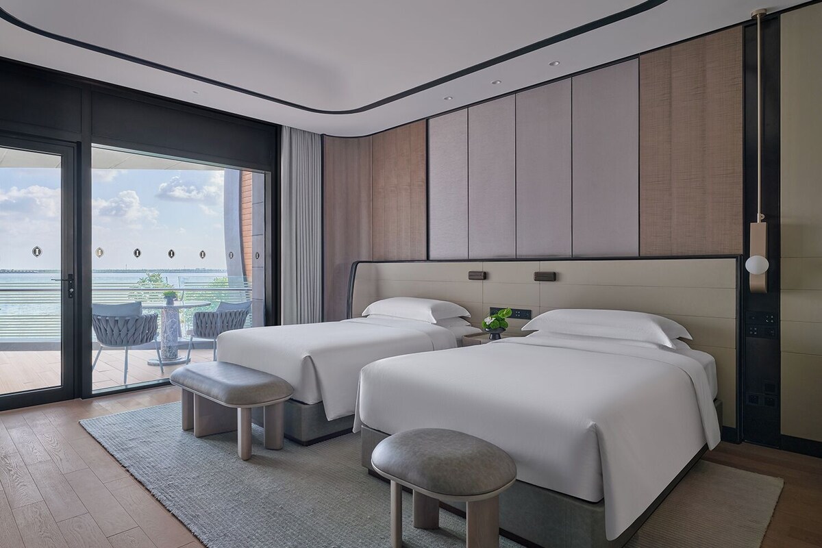 Een moderne hotelkamer met twee bedden, een grote schuifdeur naar een balkon met uitzicht op het water, en een kleine tafel met stoelen buiten.