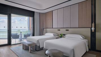 Et moderne hotelværelse med to senge, en stor skydedør til en balkon med udsigt over vandet, og et lille bord med stole udenfor.
