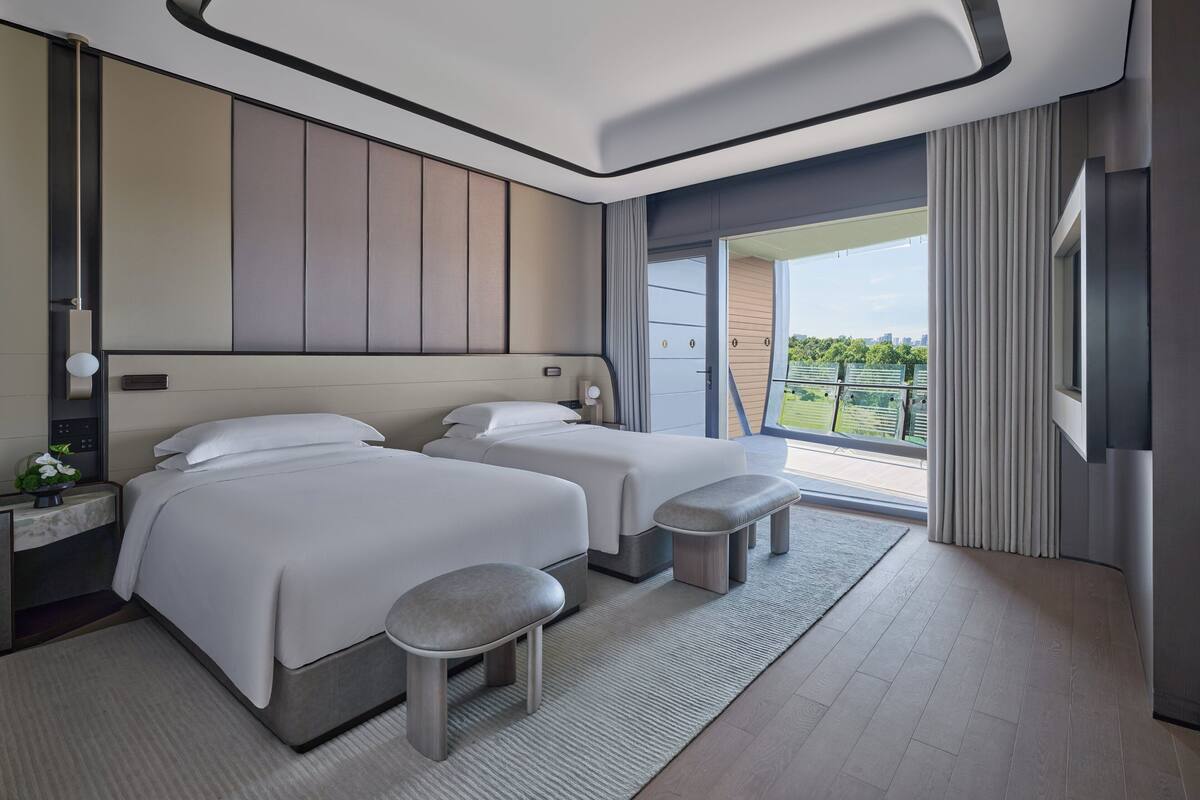 A modern hotel room with two beds, a small stool, a nightstand, and a large window offering a view of the outdoors.