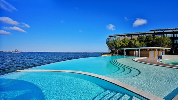 Un hotel moderno con una amplia piscina infinita con vista al mar, un área de estar cubierta y un edificio de cristal rodeado de árboles.