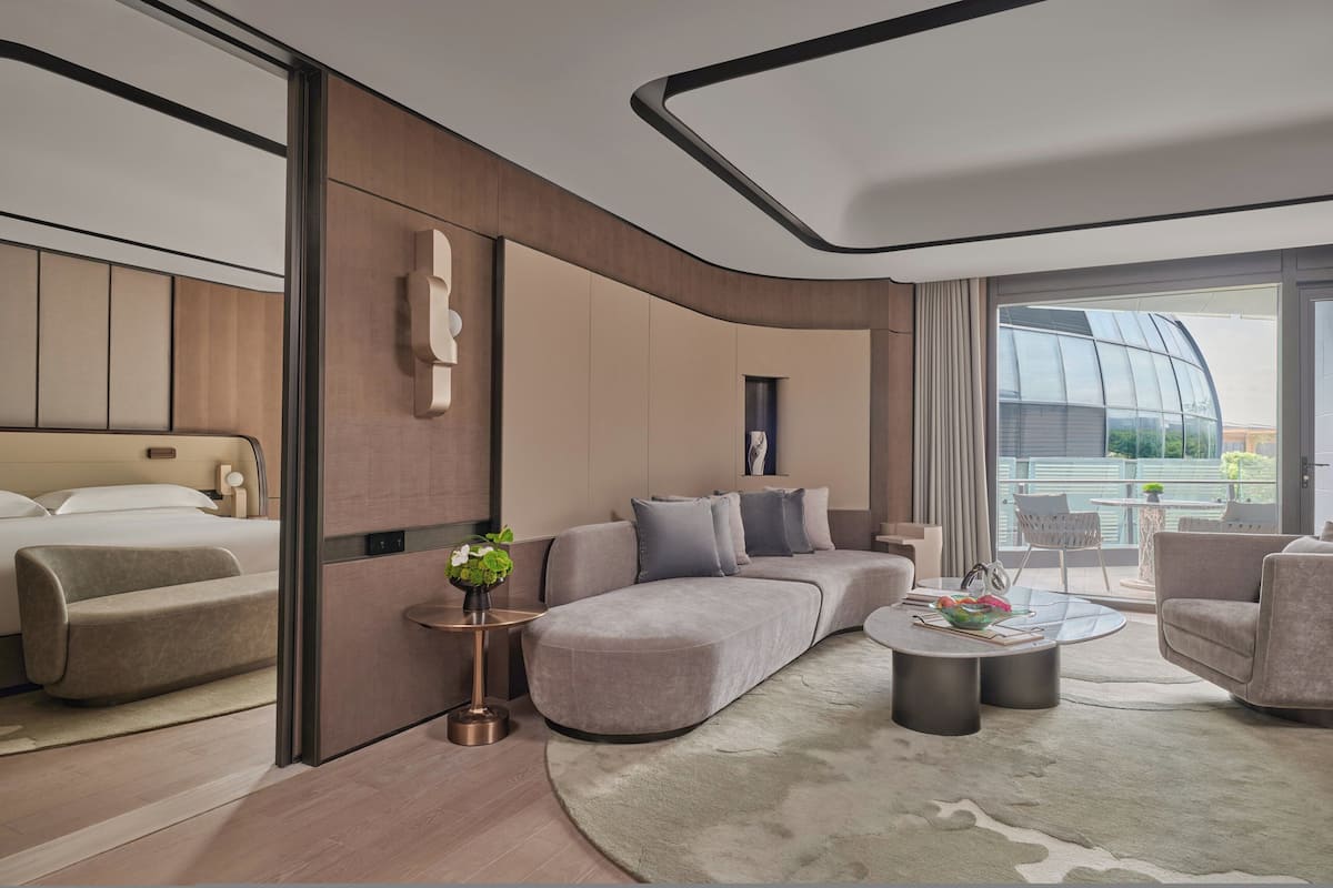 A modern hotel room with a large bed, a sofa, a coffee table, and a balcony with a view.