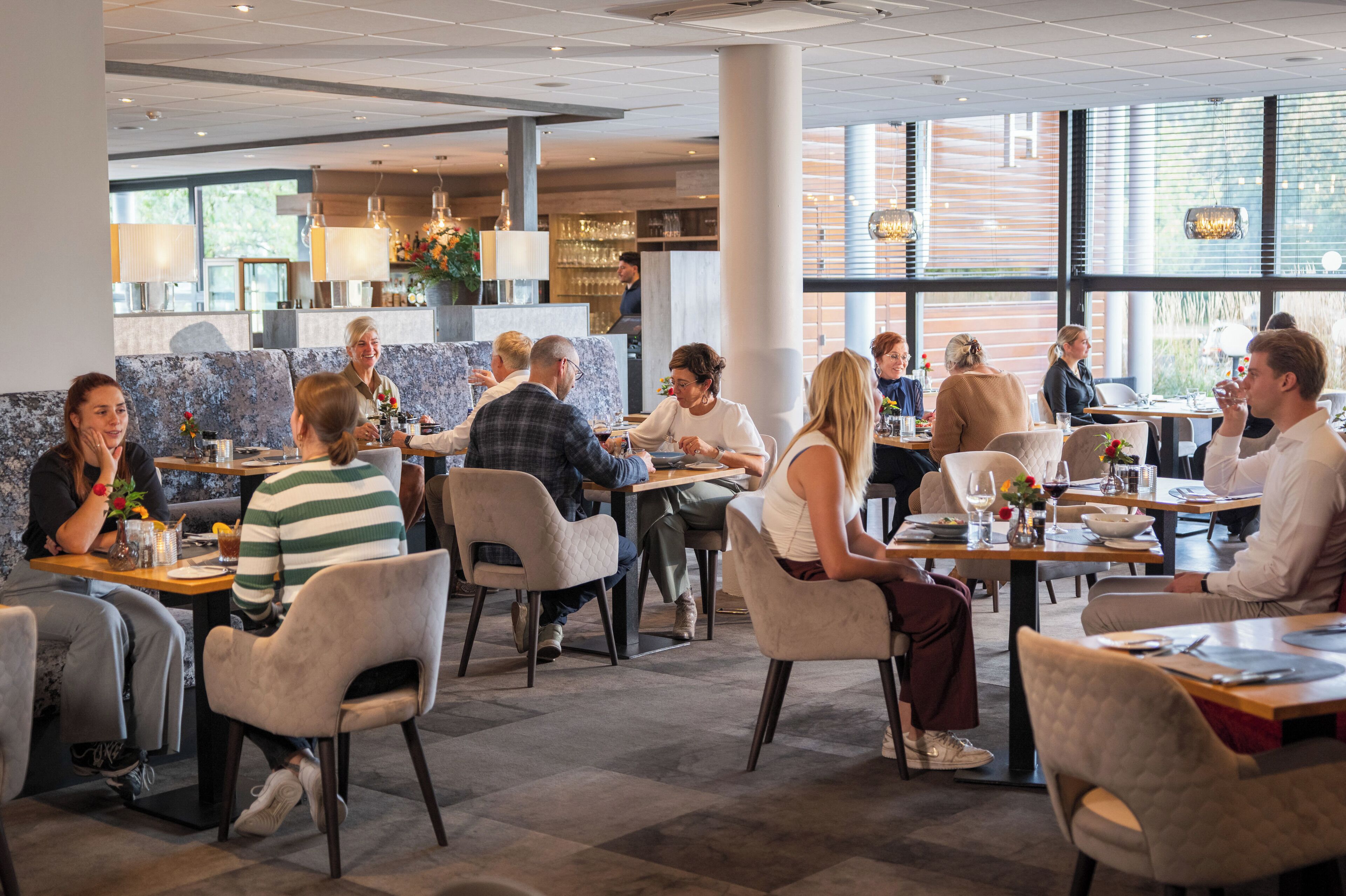 Een moderne eetruimte met meerdere tafels en stoelen, grote ramen en een centraal gelegen bar.
