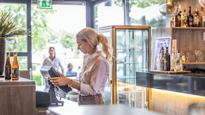 A barista at a coffee shop counter with a cash register, a potted plant, and a glass display case in the background. - Asteria Hotel Venray (Venray)