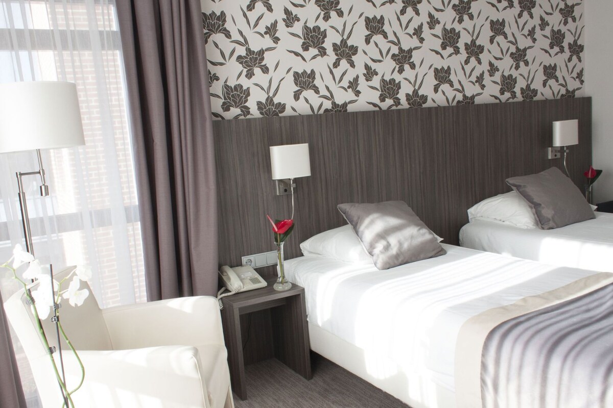 A hotel room with two beds, a chair, a nightstand with a phone, a lamp, and a vase with flowers.