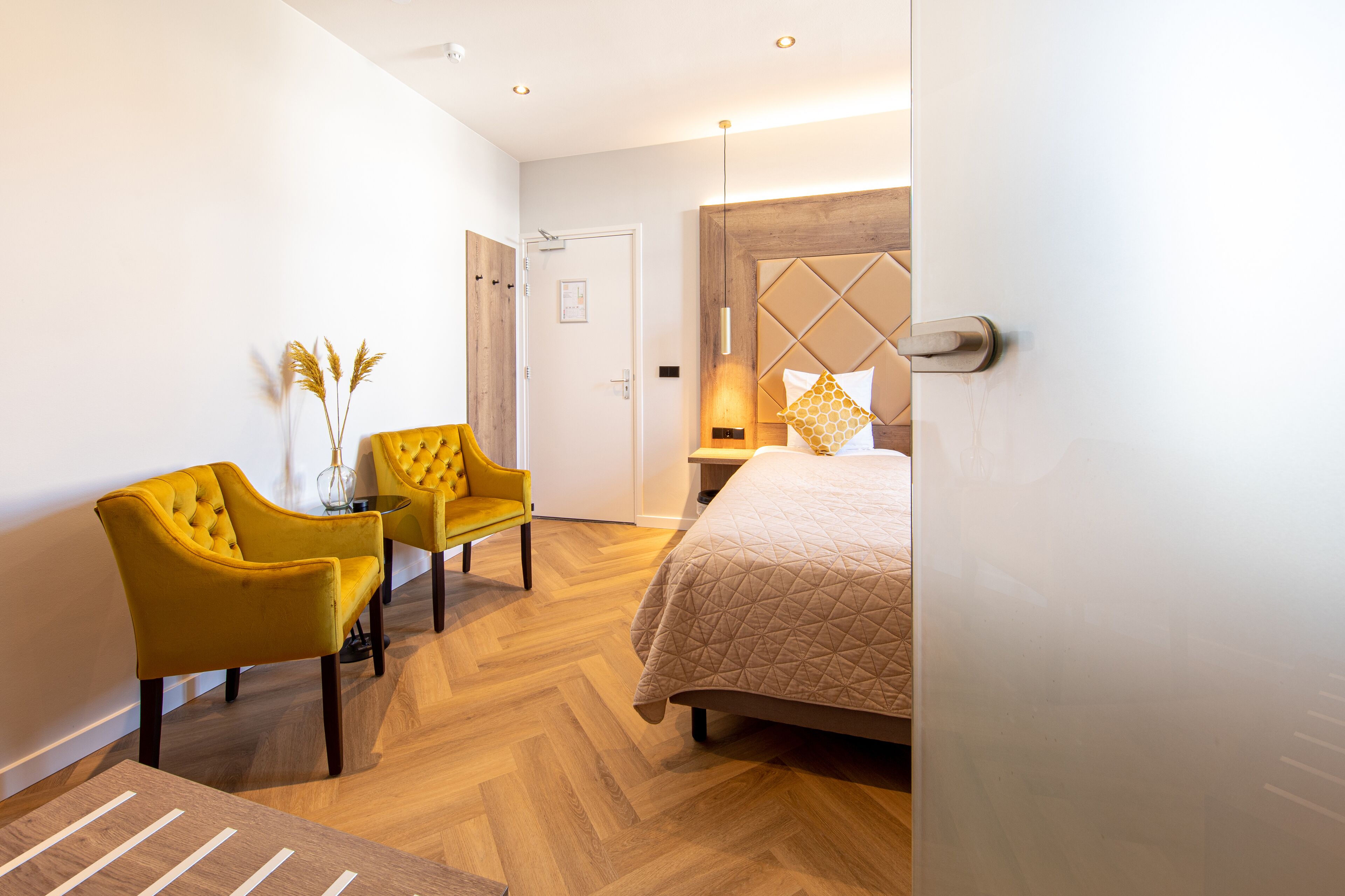 Una habitación de hotel moderna con una cama, dos sillones amarillos, una mesita con un florero y un suelo de madera con patrón de espiga.