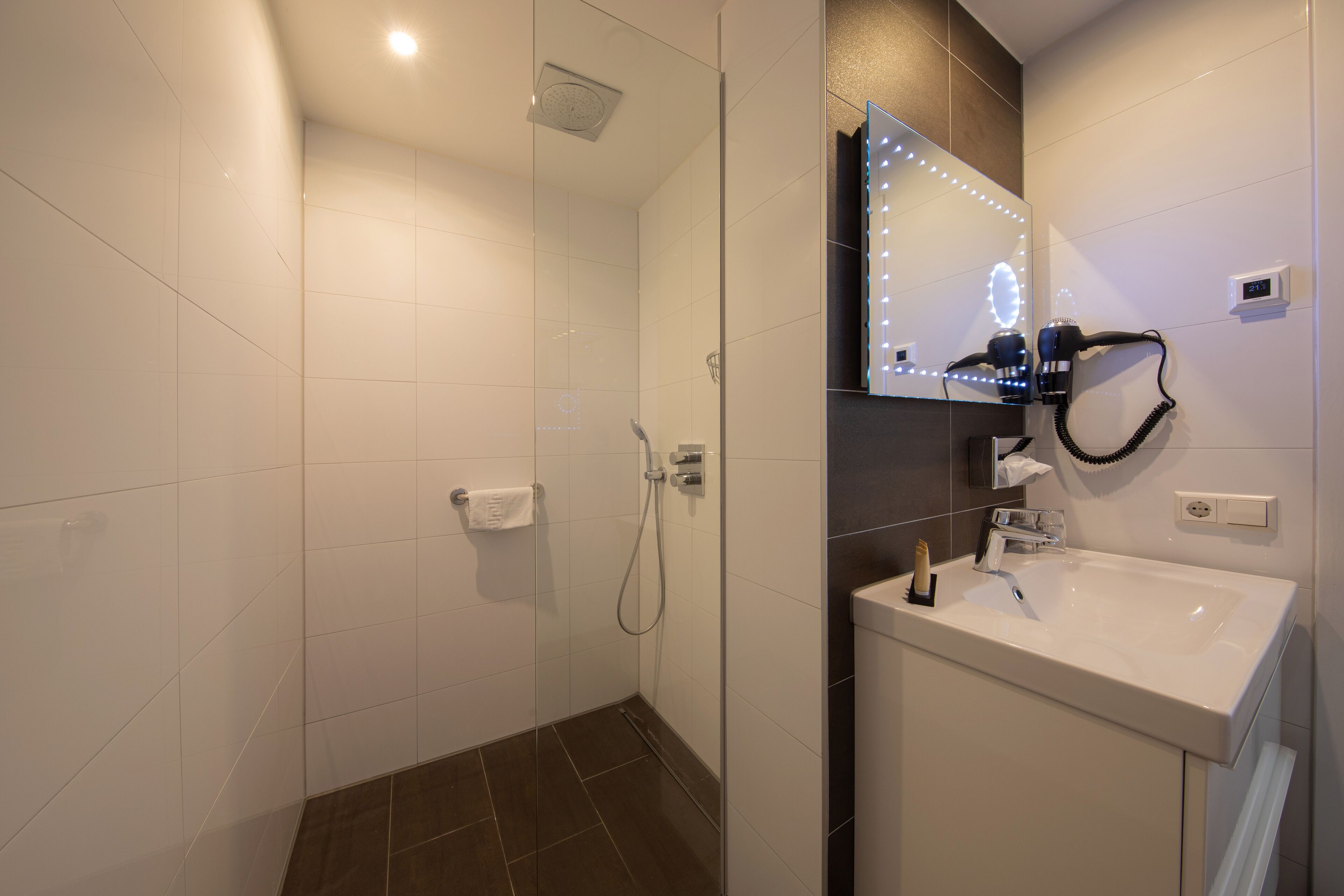 Un baño moderno con ducha envuelta en vidrio, tocador con espejo y secador de pelo en la pared.
