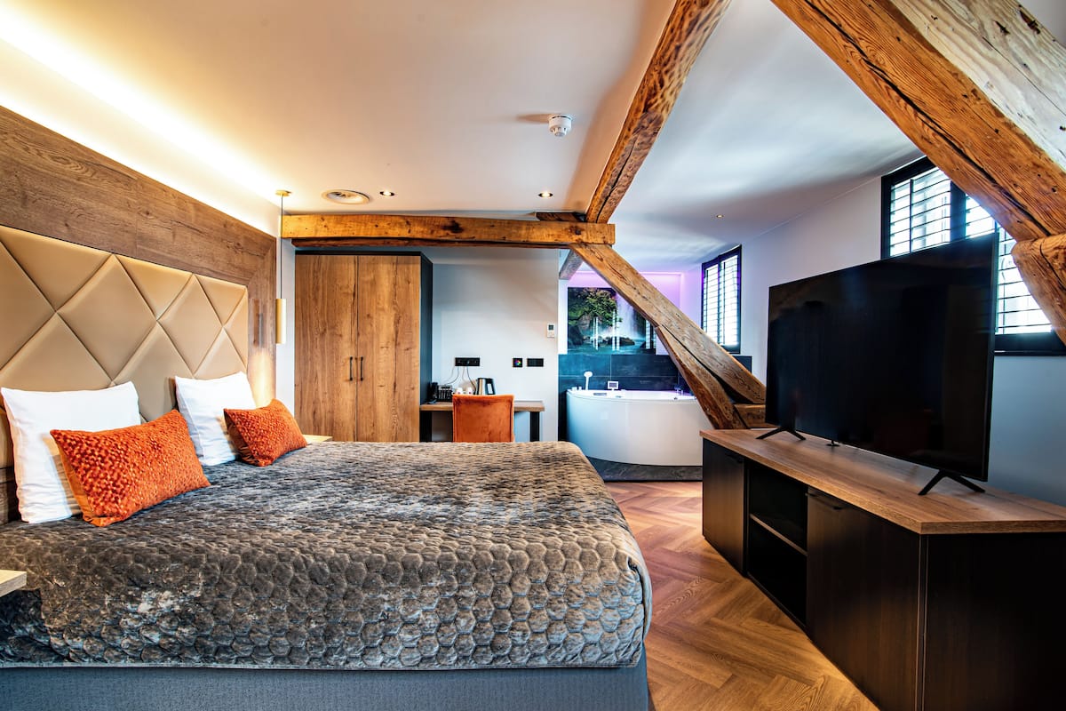 A modern bedroom with a large bed, wooden ceiling beams, a television, and a view of the outside.