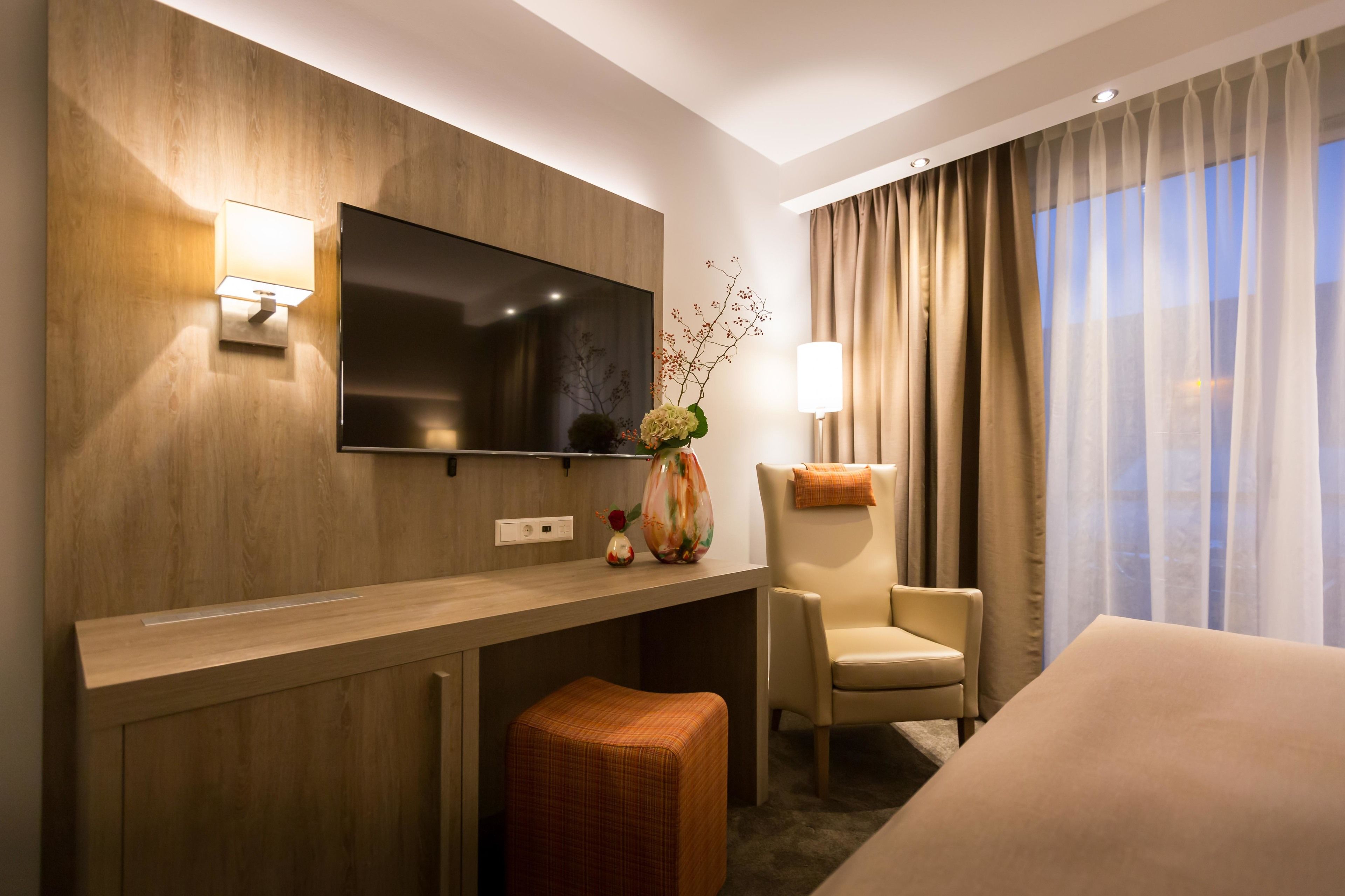 Une chambre d’hôtel dotée d’un téléviseur à écran plat fixé à un mur en bois, d’un bureau avec une lampe, d’une chaise, d’un lit et d’une fenêtre avec des rideaux.