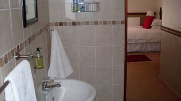 Un baño con lavamanos, espejo y artículos de aseo. Se ve una cama en la habitación contigua.
