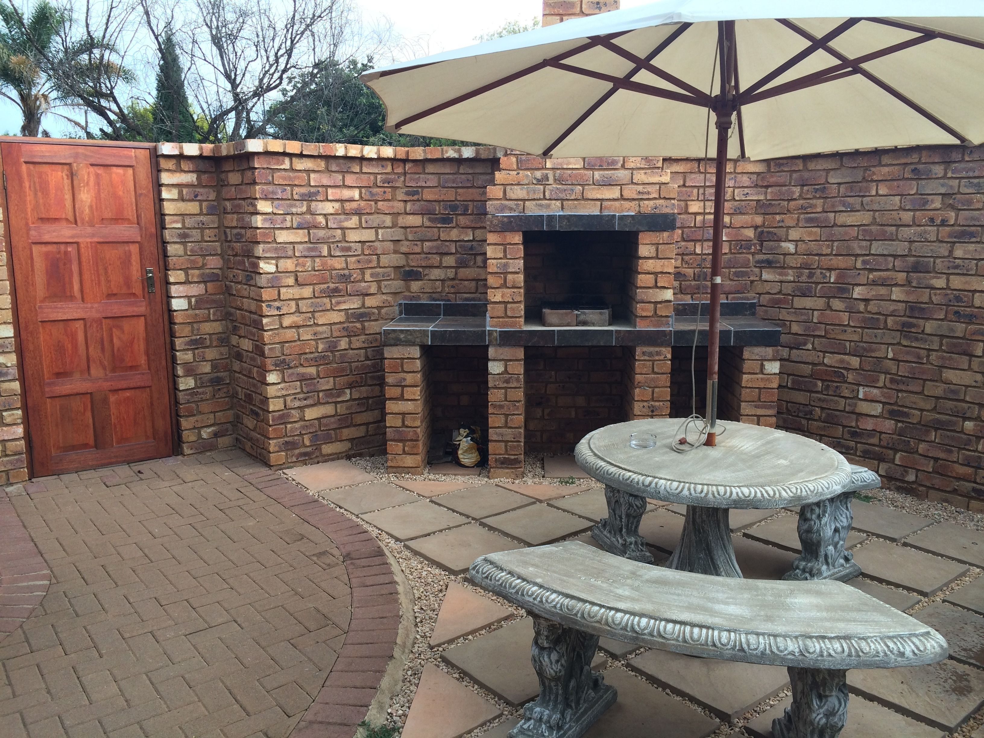 Un patio de ladrillo con puerta de madera, una parrilla y una mesa de piedra bajo una pérgola.