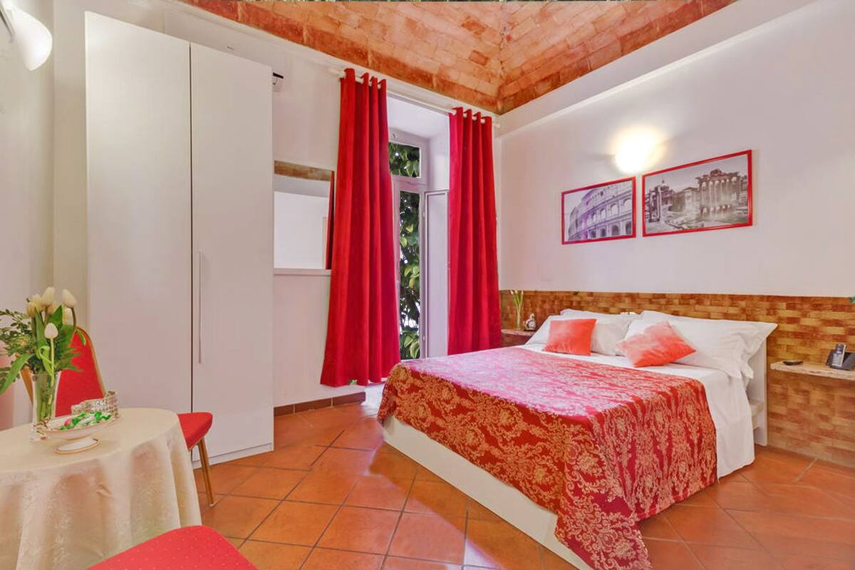 Une chambre à coucher avec un lit, des rideaux rouges, une table sur laquelle est posé un vase de fleurs, et deux tableaux encadrés accrochés au mur.