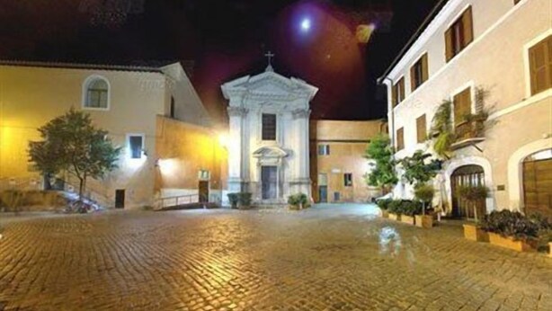 A cobblestone courtyard with a central building featuring a cross on top, flanked by two buildings with balconies. - Trastevere Relais (Rome)