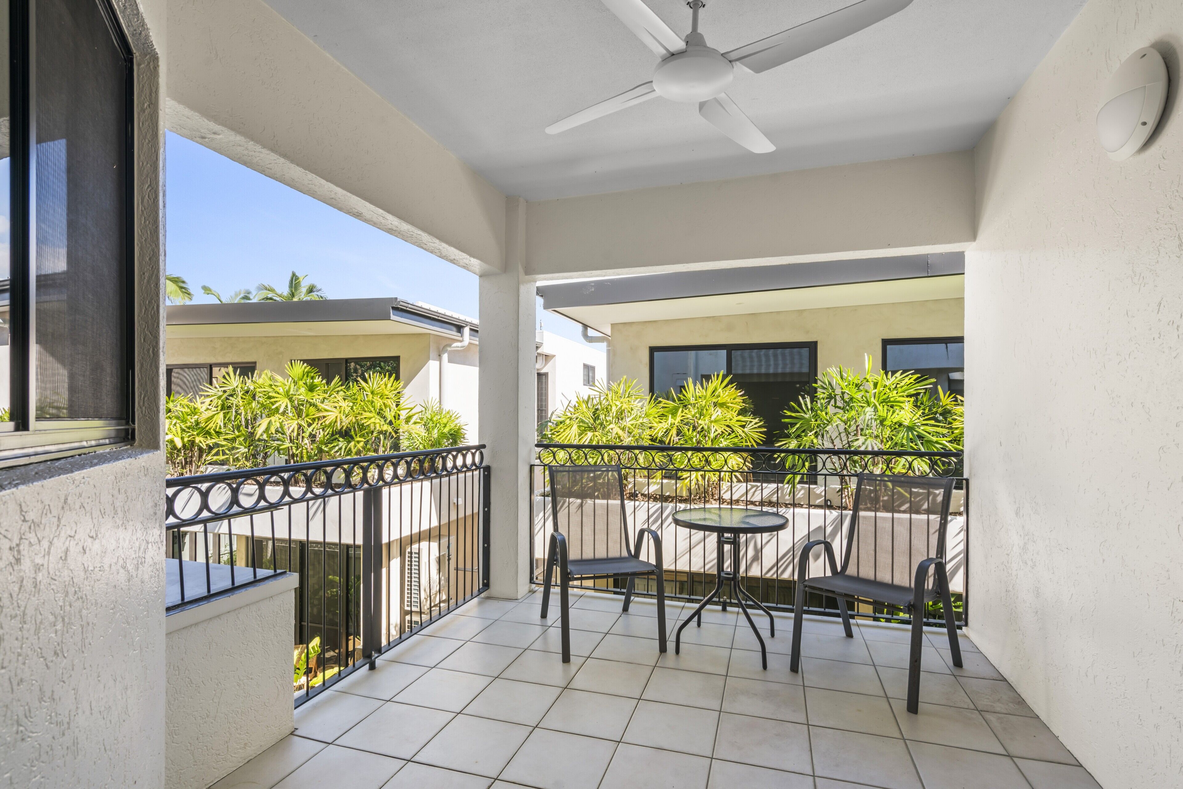 A balcony with a ceiling fan, a round table, two chairs, and a view of neighboring buildings and greenery.