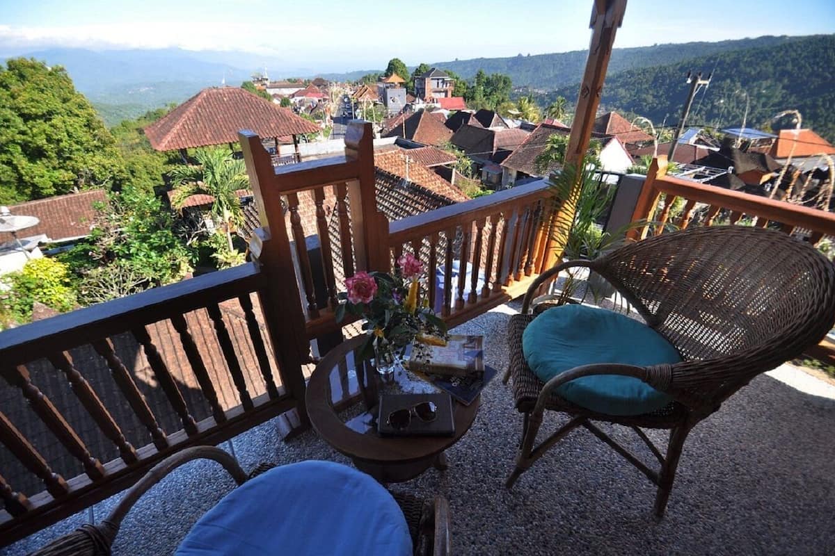 A balcony with wicker furniture, a table with a vase of flowers, and a view of a village with red-roofed houses.