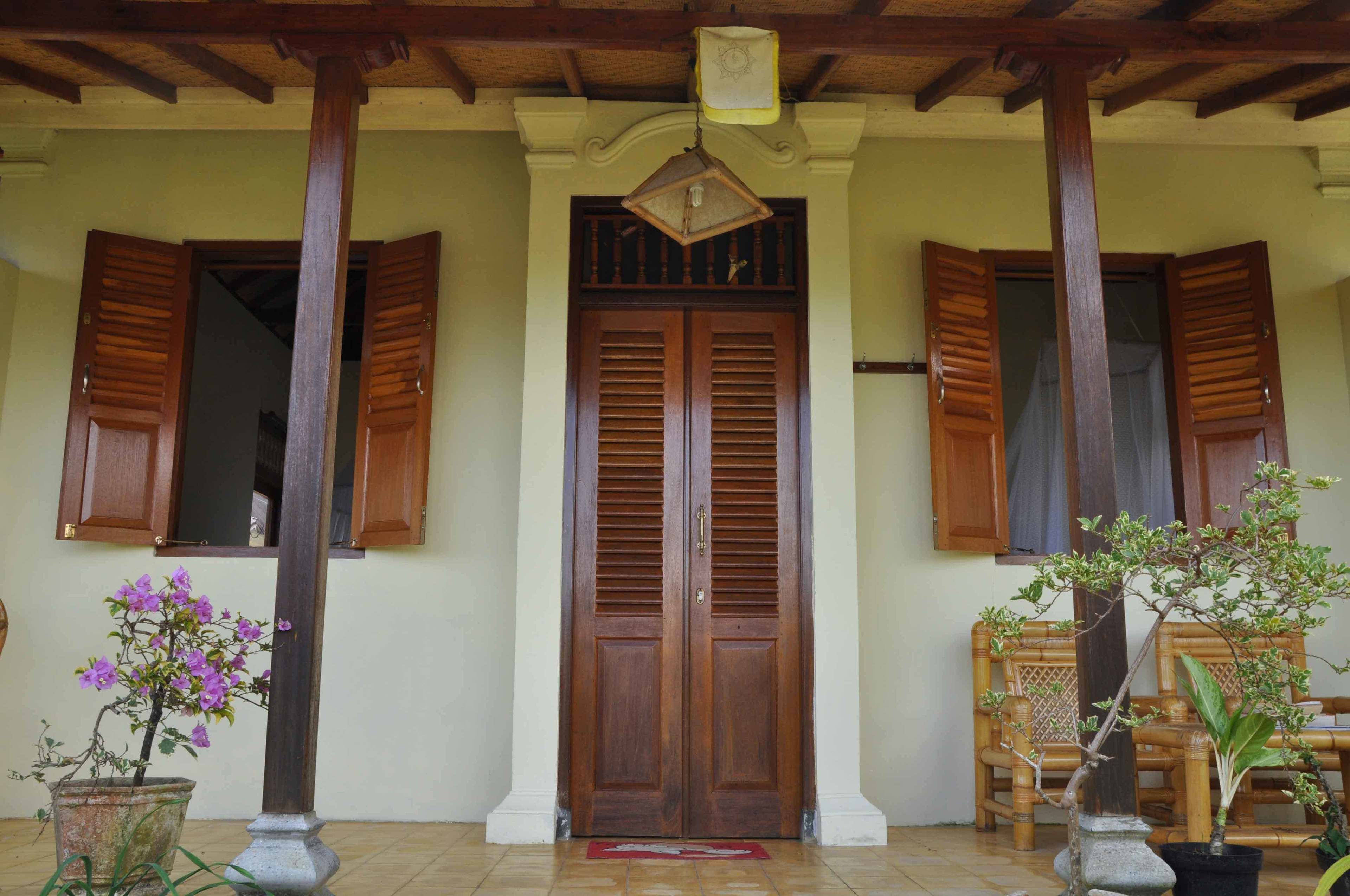 A wooden door with a window above it, flanked by wooden shutters, and a potted plant to the left.
