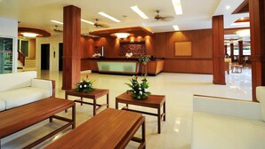 Um hall de hotel moderno com uma mesa de recepção, mobiliário de madeira e uma escadaria ao fundo.