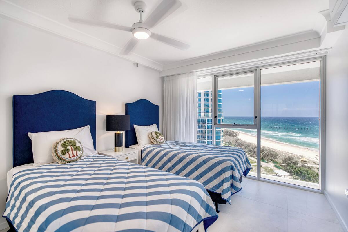 A hotel room with two beds, a ceiling fan, and a large window with a beach view.