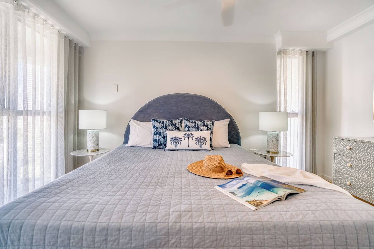 A neatly made bed with a grey quilt, two patterned pillows, a straw hat, and a magazine on the bed.