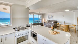 A modern kitchen with a large island, a dining area with wooden chairs, and a view of the ocean. - Talisman Beachside (Broadbeach)