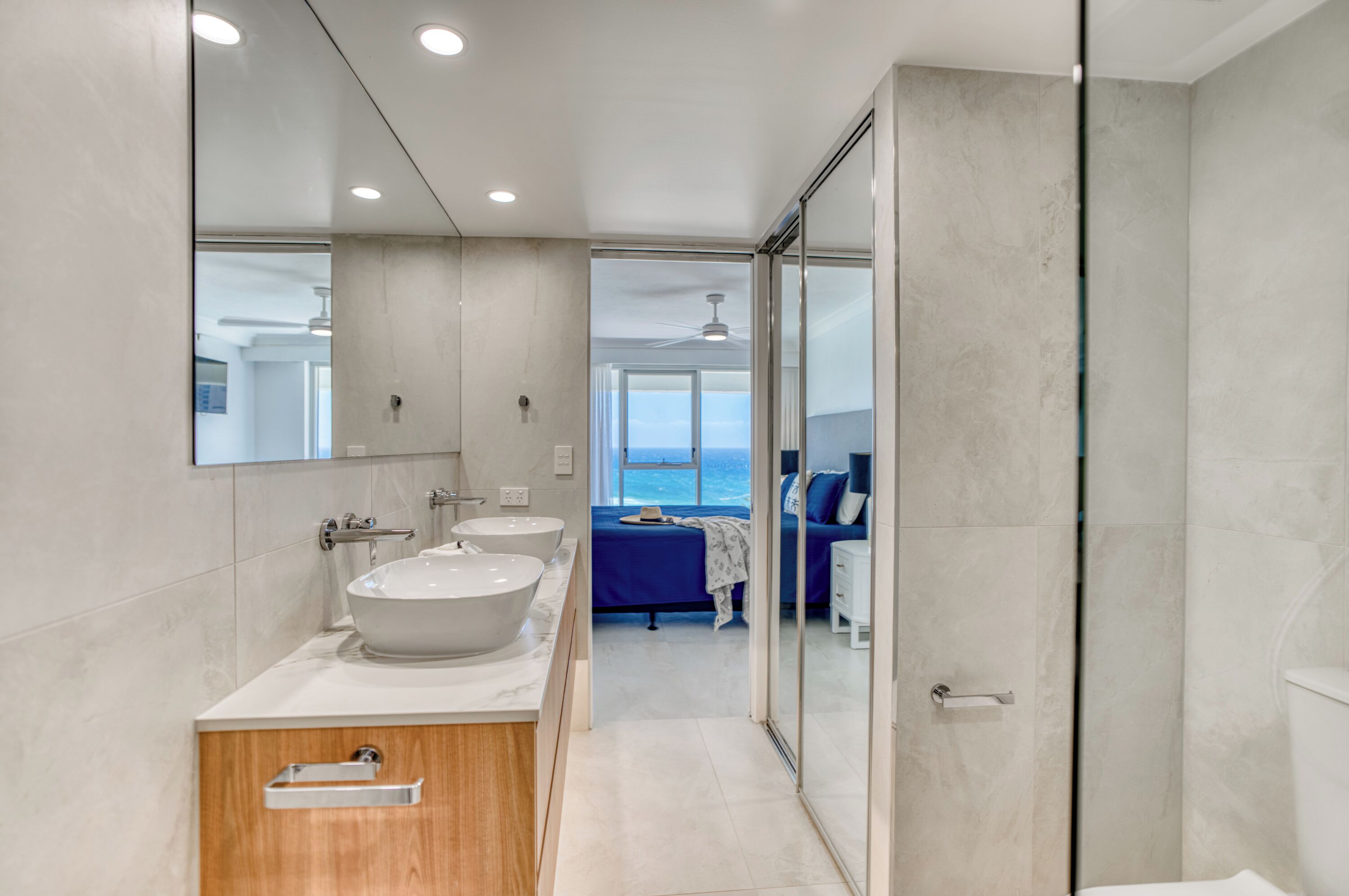 Un baño moderno con un espejo grande, dos lavamanos y vistas a un dormitorio con cama y vistas al océano.