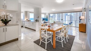 Una cocina moderna con armarios blancos, un comedor con mesa de madera y sillas blancas, y ventanas amplias que ofrecen vistas a rascacielos.