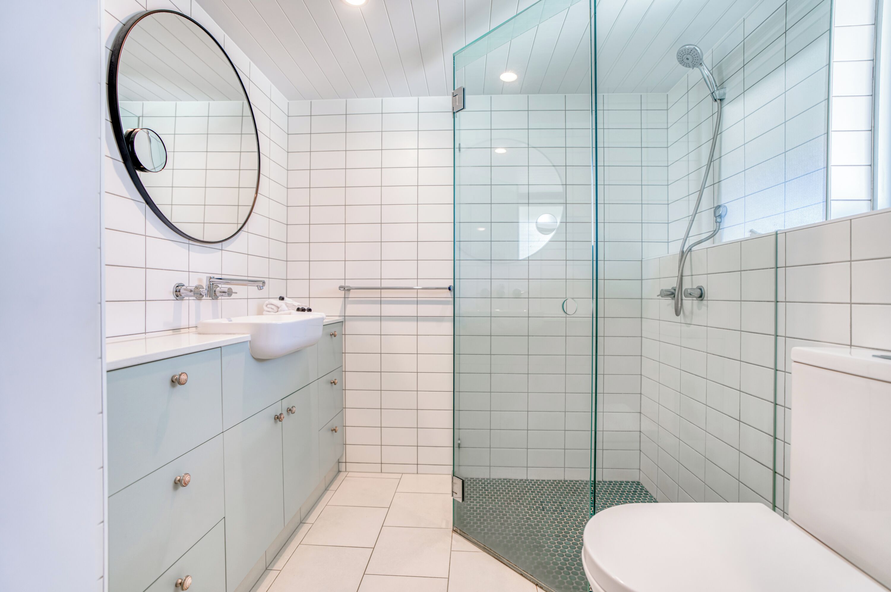 A modern bathroom with a glass-enclosed shower, a round mirror, and light blue cabinets.
