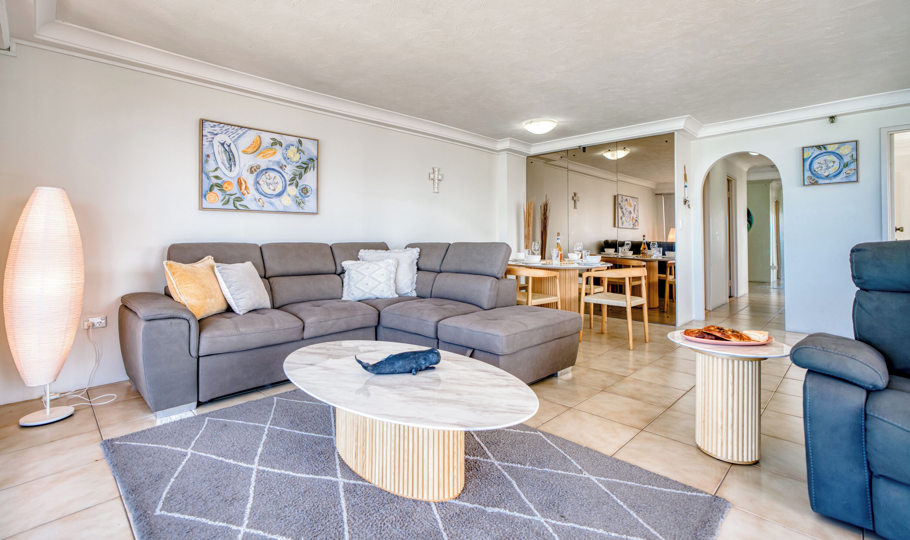 A modern living room with a grey sectional sofa, a round coffee table, and a dining area with wooden chairs.