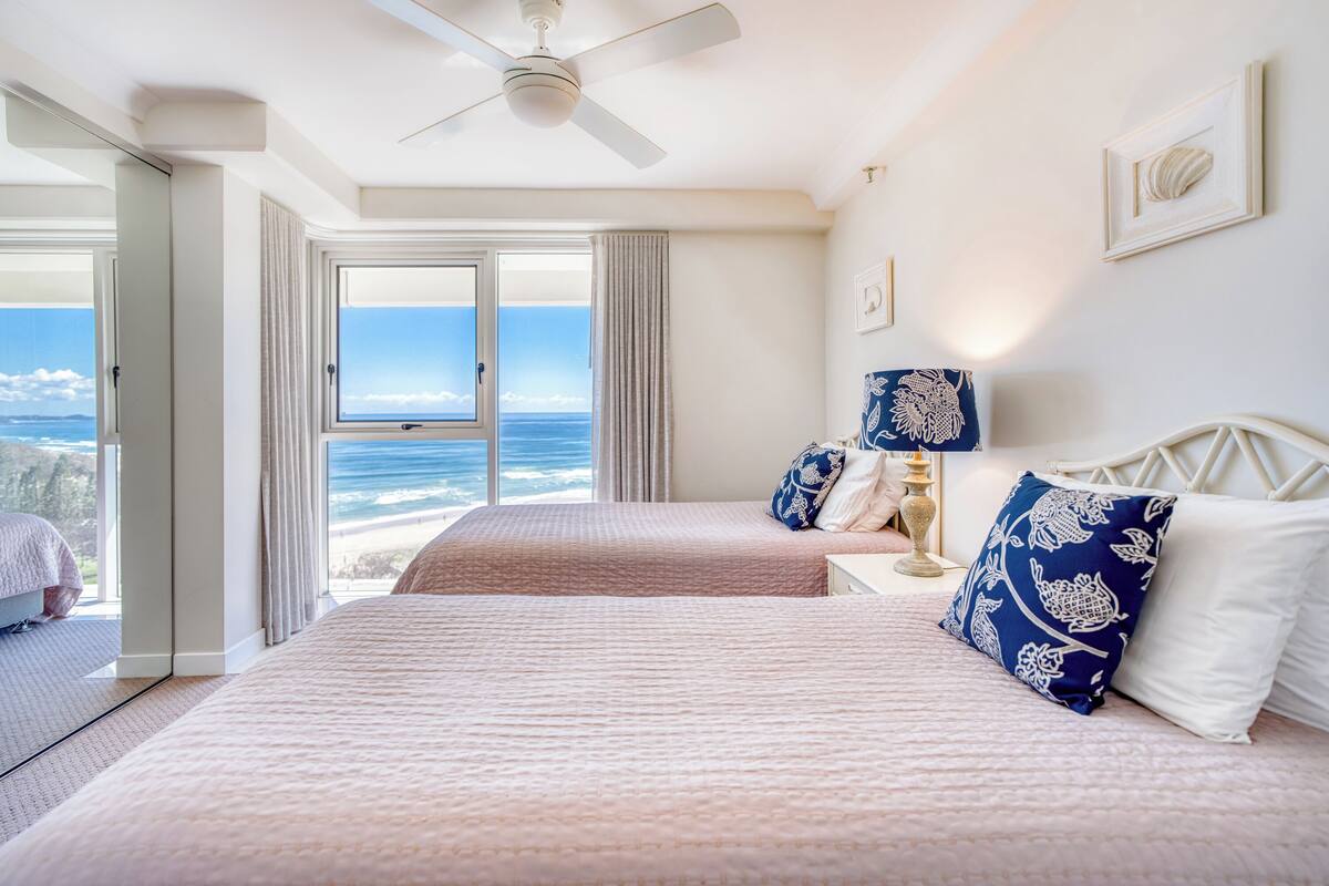 A hotel room with two beds, a ceiling fan, a nightstand with a lamp, and a large window offering a view of the ocean.