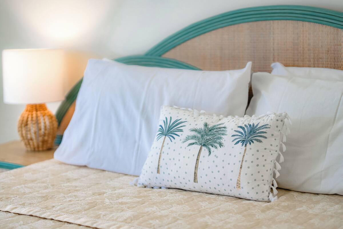 A bed with a beige quilt, white pillows, and a decorative pillow with palm tree designs.